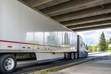 Long hauler big rig semi truck transporting cargo in dry van semi trailer running on the road under the bridge