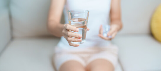 woman hand holding medicine painkiller pill and water glass on the sofa at home, taking for...