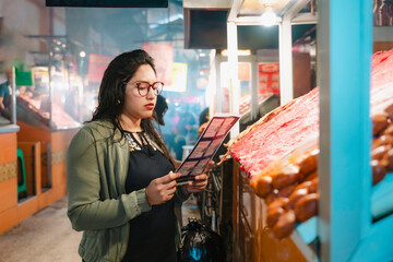 Young Latina woman reading a menu at a market in Oaxaca, Mexico, deciding on local food to try, with a focused expression.