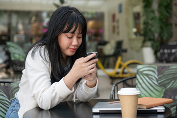 A young, happy Asian woman using her smartphone while sitting at an outdoor table of a cafe.