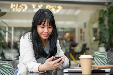 Fototapeta premium A young, happy Asian woman using her smartphone while sitting at an outdoor table of a cafe.
