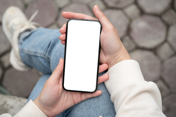 A woman holding a smartphone with a white-screen mockup over her lap while sitting outdoors.