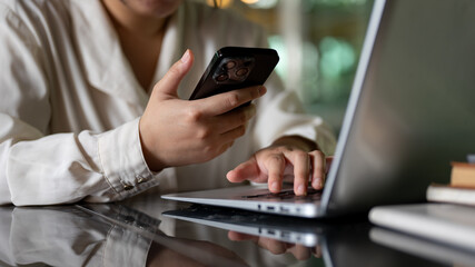 A cropped image of a businesswoman is multitasking, using her smartphone and laptop computer.