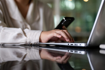 A cropped image of a businesswoman is multitasking, using her smartphone and laptop computer.