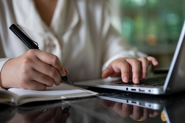 A cropped image of a businesswoman taking notes in her notebook while working on her laptop computer
