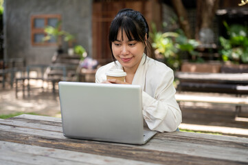 A businesswoman sipping coffee while focusing on reading emails on her laptop computer in a garden.