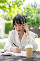 A focused Asian woman is talking on the phone with her colleague while working remotely in a garden.