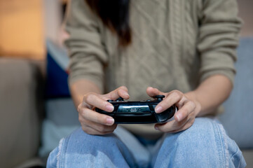 A close-up image of a woman is holding a joystick and playing a video game on the sofa at home.