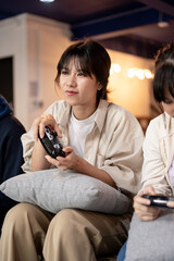 Fototapeta premium A young, beautiful Asian girl is enjoying playing a video game on a couch at home with her friends.