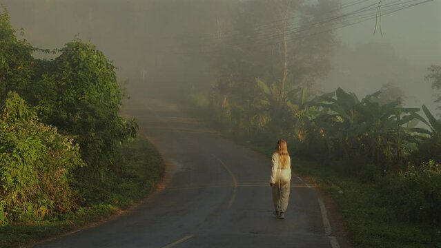 Lonely woman walking down misty, secluded road during early morning hours. road is surrounded by lush green vegetation and tall trees, creating serene and peaceful atmosphere.