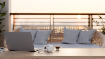 A computer, headphones, and a coffee cup on a wooden coffee table on a balcony with a wicker couch.