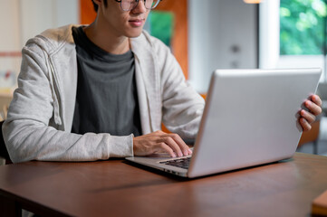 A close-up image of an Asian man working on his laptop computer and working at a table indoors.