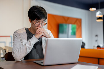 A thoughtful young Asian man is working on his laptop, looking at the screen with a serious face.
