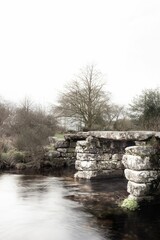 A serene winter landscape featuring an old stone bridge over a calm river with bare trees