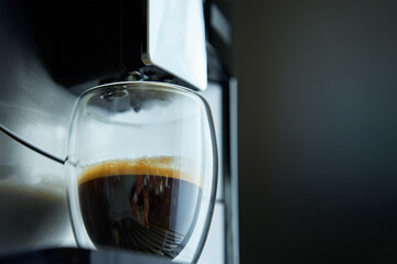 Fresh espresso in clear glass at morning. Coffee machine on dark background, close up. Modern coffee maker with freshly brewed coffee in glass cup. Kitchen appliances