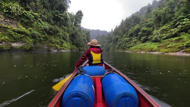 Woman in a red jacket, hat, and yellow life vest quickly paddles down the Whanganui River in her canoe between two forest cliffsides