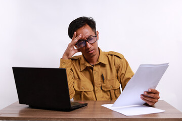 Stressed male teacher in civil servant uniform with paper and laptop on the desk holding a paper