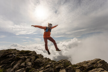 A man is jumping in the air on a mountain. The sky is cloudy and the sun is shining
