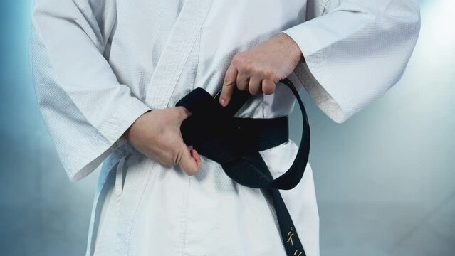 Karate fighter ties black belt before training. Man tying belt on kimono. Fighter preparing for karate workout in practice room. Sport concept. Close-up in 4K, UHD