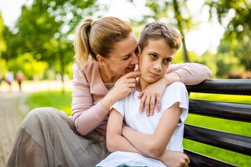 Naklejka premium Happy mother is sitting with her son on bench in park. Boy is offended and pouting.