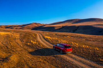 A red SUV drives along a winding dirt road through a beautiful landscape of rolling hills and golden grasses in Gareji area in Georgia. The car travel towards horizon, creating a sense of adventure