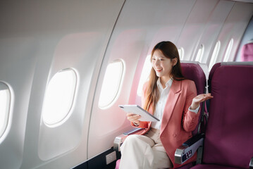 Portrait of a successful Asian businesswoman or entrepreneur in a formal suit on an airplane sitting in business class using a phone, computer laptop. Travel in style, work