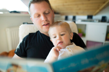 Father holding his son in arms and reading book together. Dad spending time with toddler in his room. Unconditional paternal love, Father's Day.