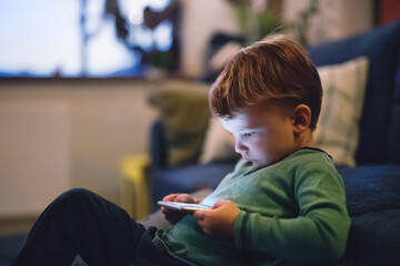 Cute boy sitting on bed watching something on smartphone. Children's screen time.