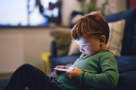 Cute boy sitting on bed watching something on smartphone. Children's screen time.