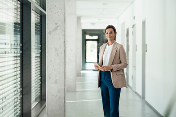Cheerful businesswoman holding smart phone and standing at office