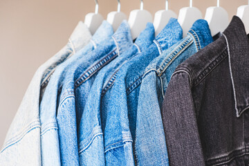 Many jean jackets on hangers, close-up view. Different shades of denim fabric colors.