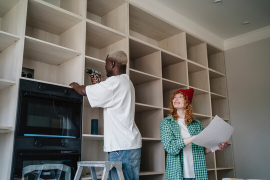 Smiling woman standing with instruction manual near man using drill on rack at home