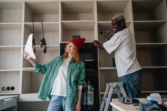 Smiling woman reading instruction manual with friend using drill on cabinet at home