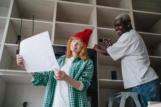 Smiling young woman reading instruction manual with friend using drill on cabinet at home