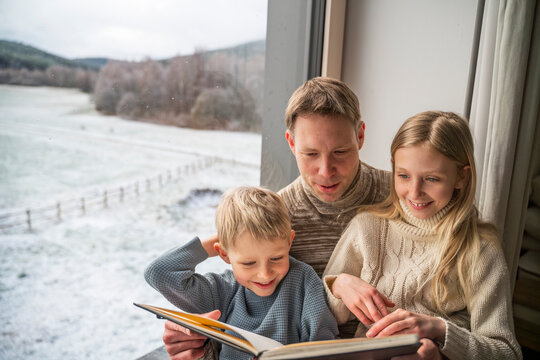 Father with children reading book by window at home in winter