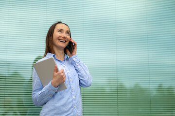 Smiling businesswoman talking on smart phone and holding tablet PC in front of glass wall