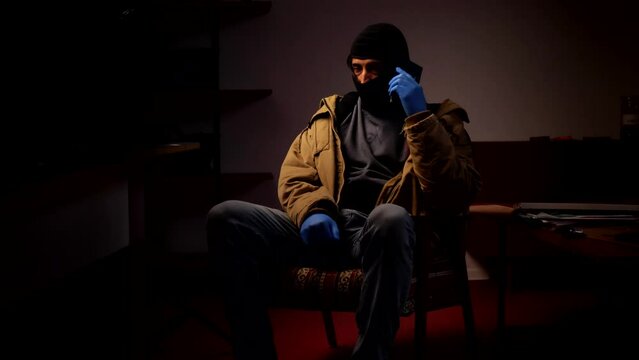 A young adult man wearing a balaclava and rubber gloves sits in a dark room and talks on the phone. Crime. Burglar