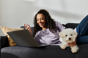 Young woman relaxing on couch with laptop, bichon frise by her side.