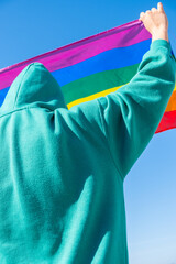 back view of a hooded person holding a rainbow-colored flag in support of the LGBT community.
