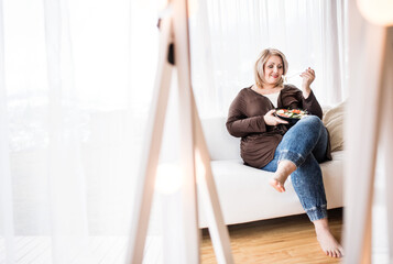 Portrait of beautiful young overweight woman at home, choosing healthy food, eating vegetable salad.