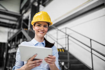 Female project manager standing in modern industrial factory, holding tablet. Manufacturing facility with robotics, robotic arms and automation. Storing products and materials in warehouse.