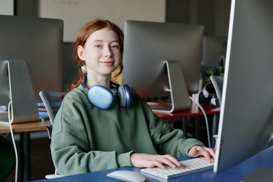 Medium portrait of cheerful teen girl wearing headphones sitting at desk in modern classroom texting on computer and looking at camera