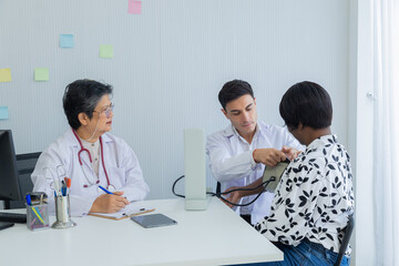 Young Caucasian doctor taking blood pressure of African American woman in clinic. Asian senior female specialist observation. middle-aged patient waiting for diagnosis of disease, medical health care
