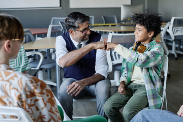 Cheerful mature school psychologist and African American teen boy doing fist bump during group...