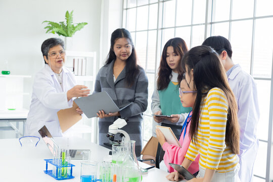 Senior Asian female teacher or scientist teaching group of diverse students in laboratory. young children using tablets and laptops, listening attentively. chemical test tubes and microscope in lab