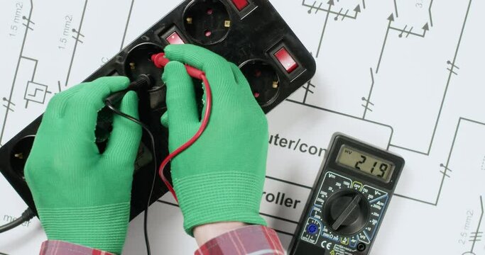 Electrician checks voltage with multimeter and puts warning sign extension sockets on drawing upper view. Worker controls safety of electrical network