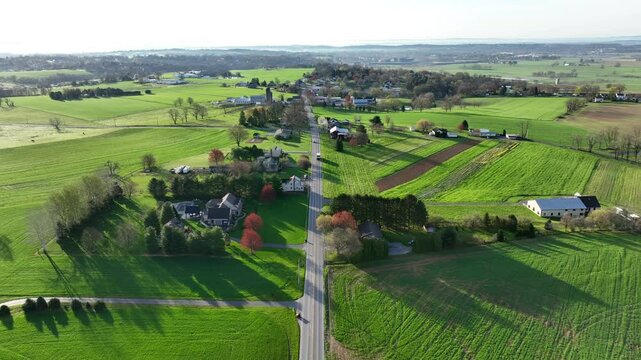 Driving cars on rural intersection in countryside of american town. SUnny day in spring season. Green farm fields and farm houses. Aerial birds eye forward shot.