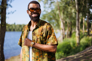 Naklejka premium Cheerful african american man in sunglasses and shirt relaxing near lake in park