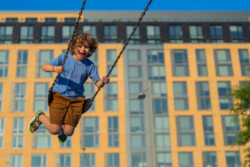 Kid swinging on chain swing on city kids playground. Swing ride. Cute child having fun on a swing on summer sky background. Blonde little boy swings at kid playground. Child swinging high.