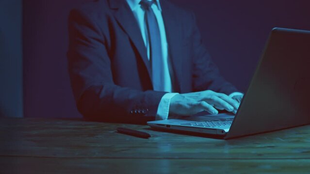 A man in a business suit with a tie works at a table with a laptop in the evening. Dark blue scheme. The hand draws an imaginary exponent curve.
